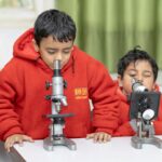 Two children in red hoodies using microscopes in a classroom setting, focusing on scientific exploration.