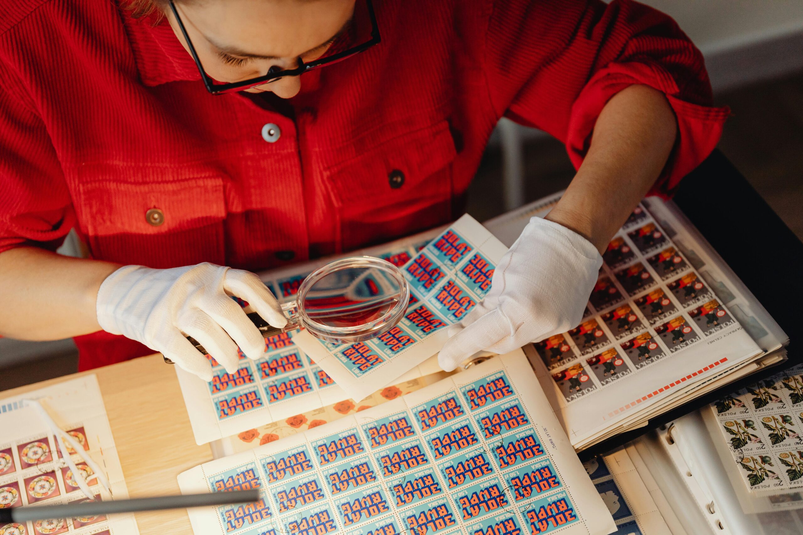 A woman wearing gloves examines stamps using a magnifying glass at a table, showcasing a philatelic hobby.