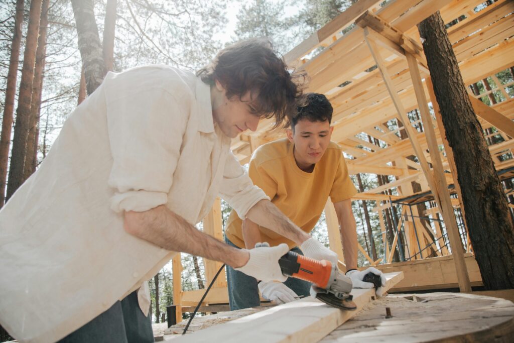 Two young men working with power tools on a woodworking project outdoors.